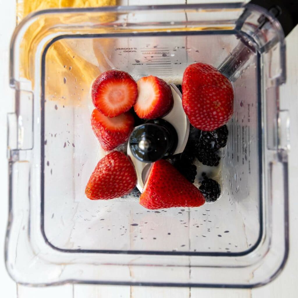 Overhead view of a blender containing whole strawberries and blackberries on a white wooden surface.
