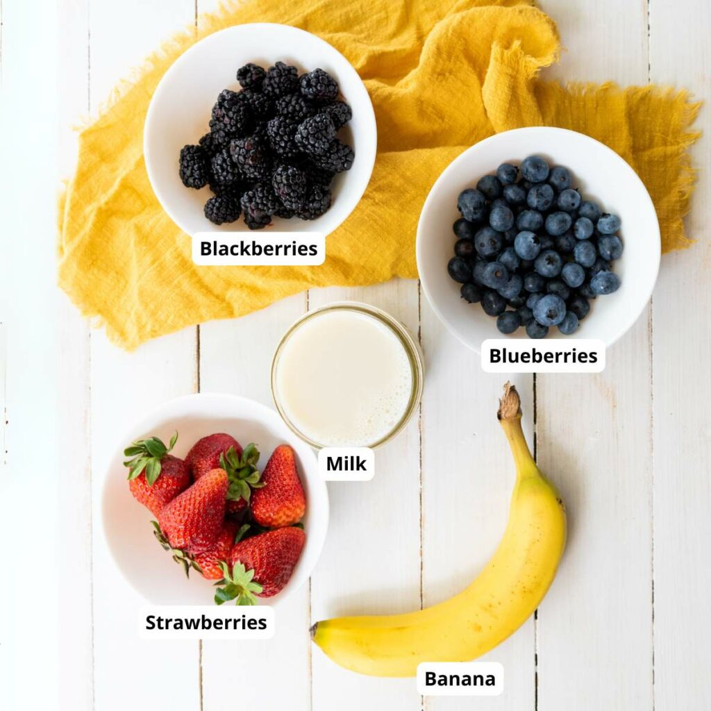 Labeled bowls and items on a white surface: blackberries, blueberries, strawberries, a glass of milk, and a banana.