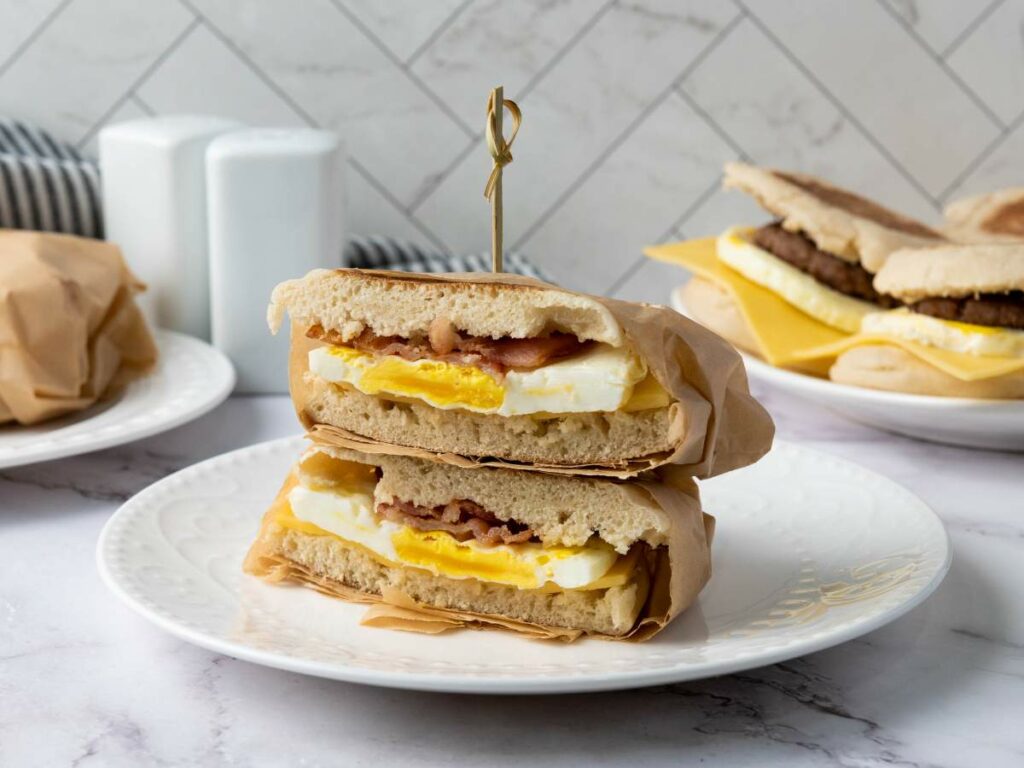 A breakfast sandwich with egg, cheese, and meat is cut in half and stacked on a white plate, with more sandwiches in the background.