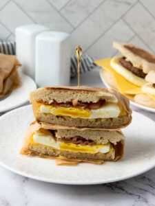 A breakfast sandwich with egg, cheese, and bacon is sliced in half and stacked on a white plate, held together with a toothpick.