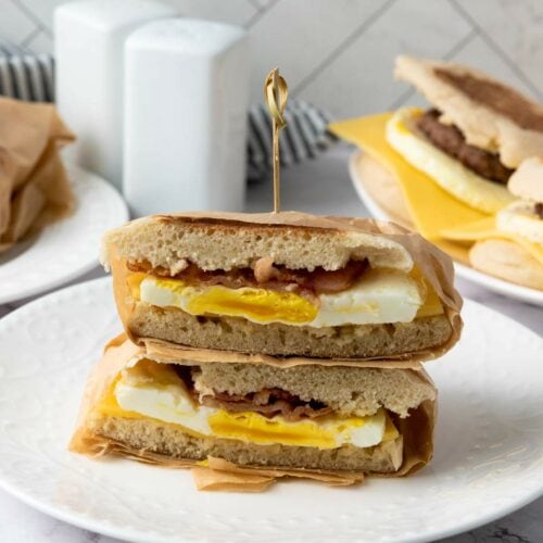 A breakfast sandwich with egg, cheese, and bacon is sliced in half and stacked on a white plate, held together with a toothpick.