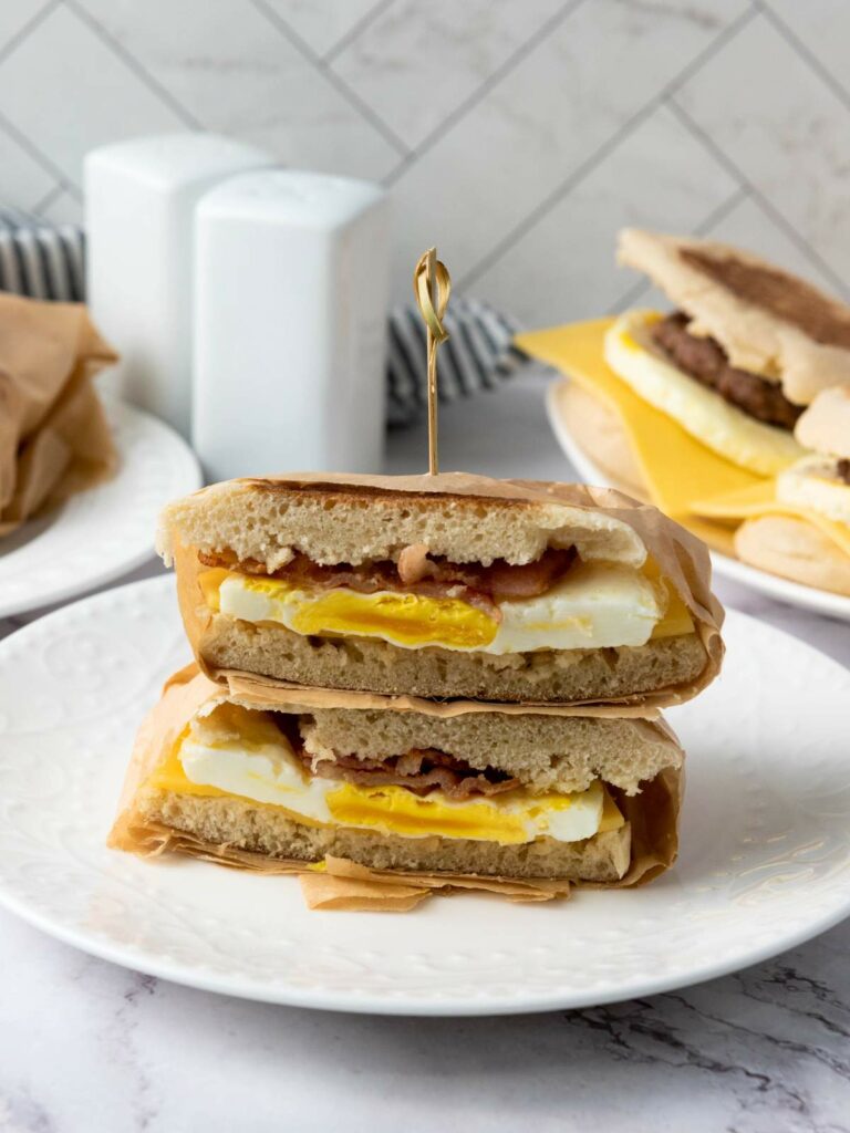 A breakfast sandwich with egg, cheese, and bacon is sliced in half and stacked on a white plate, held together with a toothpick.
