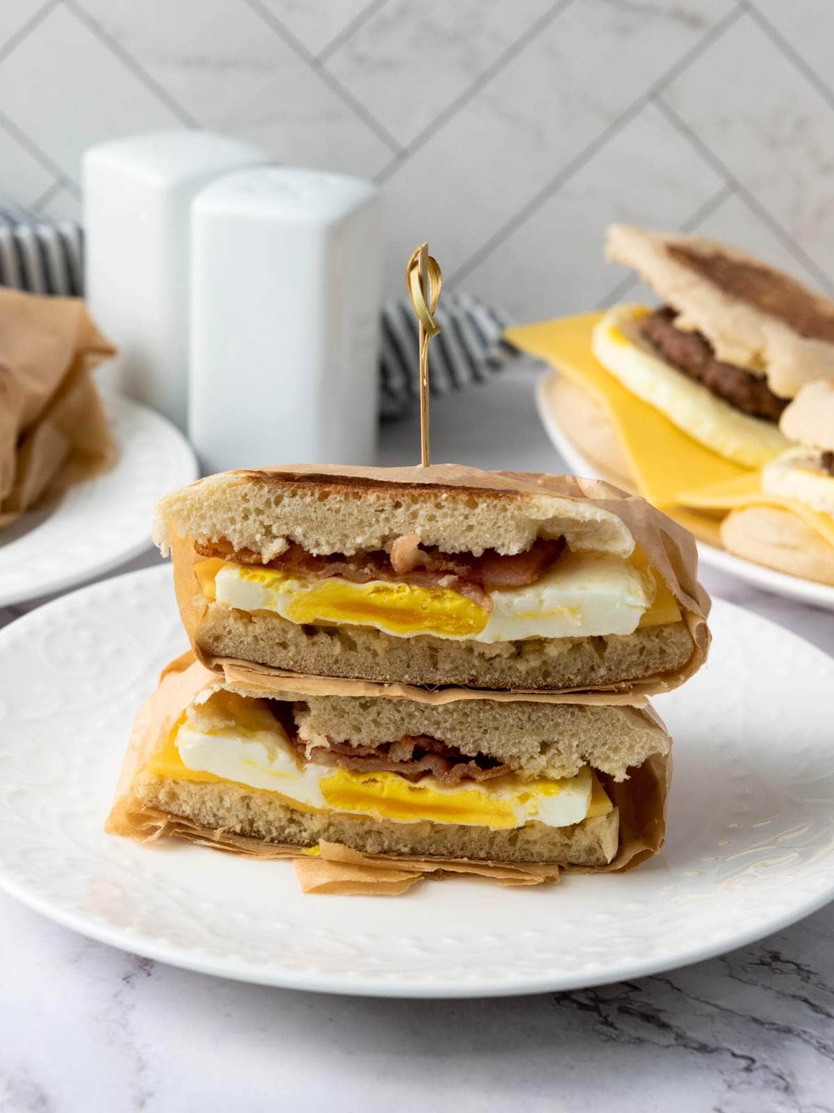 A breakfast sandwich with egg, cheese, and bacon is sliced in half and stacked on a white plate, held together with a toothpick.