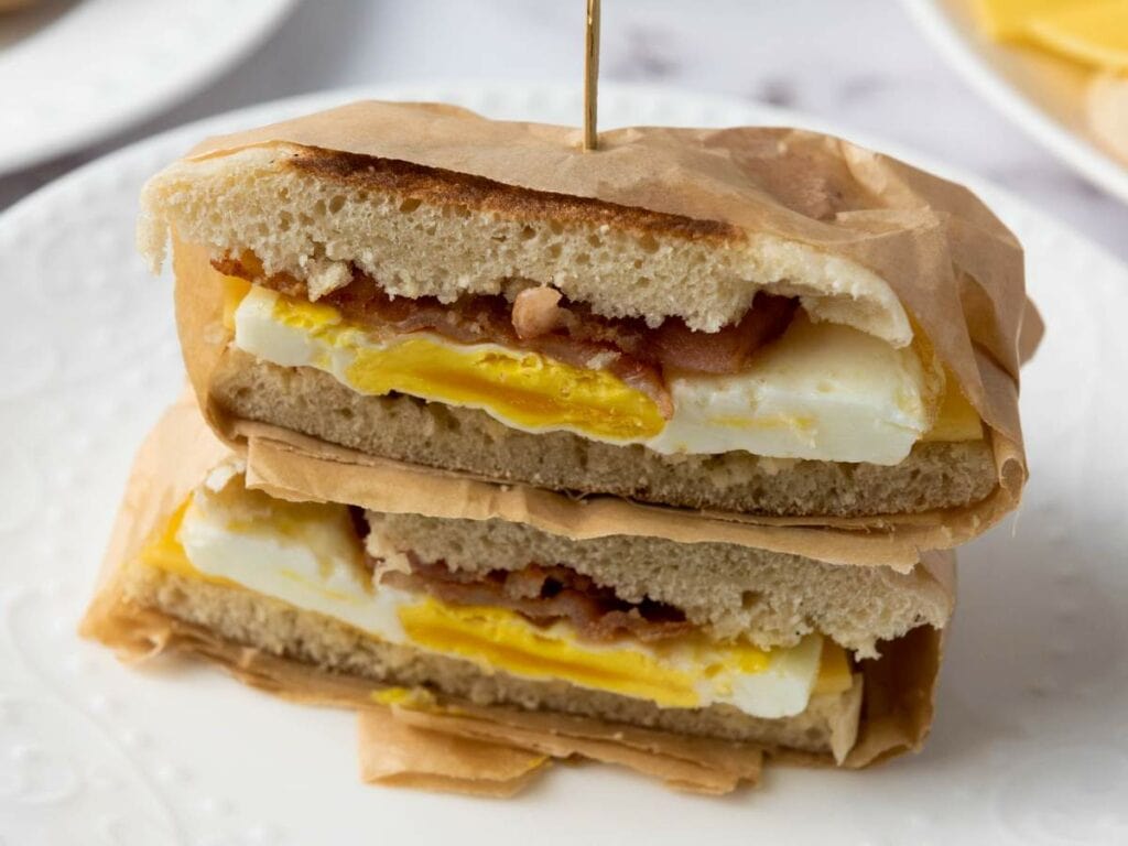 Two halves of a sandwich with egg and bacon are stacked on a white plate, wrapped partially in brown paper and secured with a toothpick.