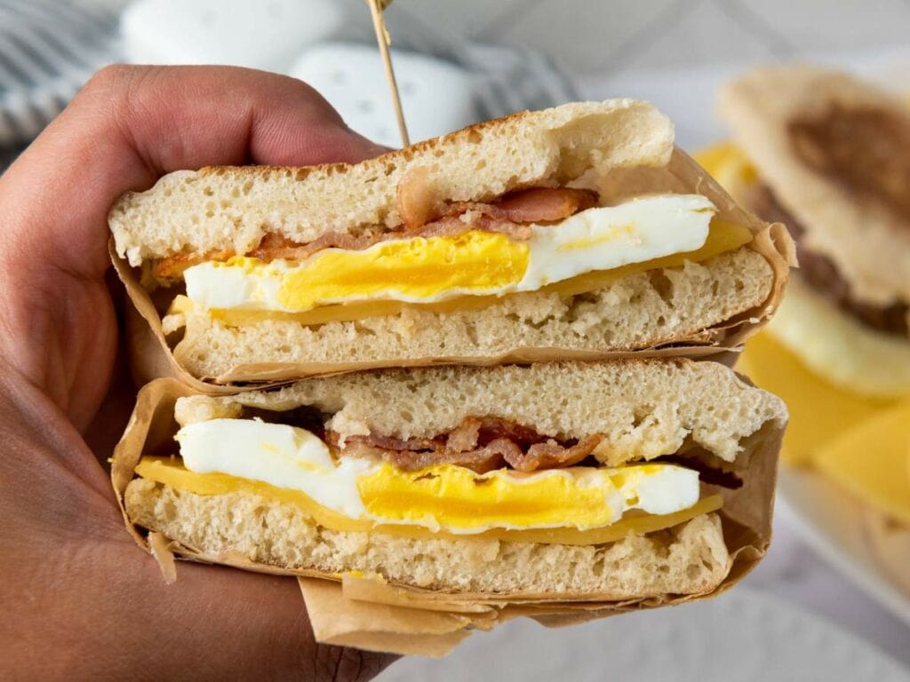 Hand holding a stacked breakfast sandwich with bacon, egg, and cheese between two halves of ciabatta bread.