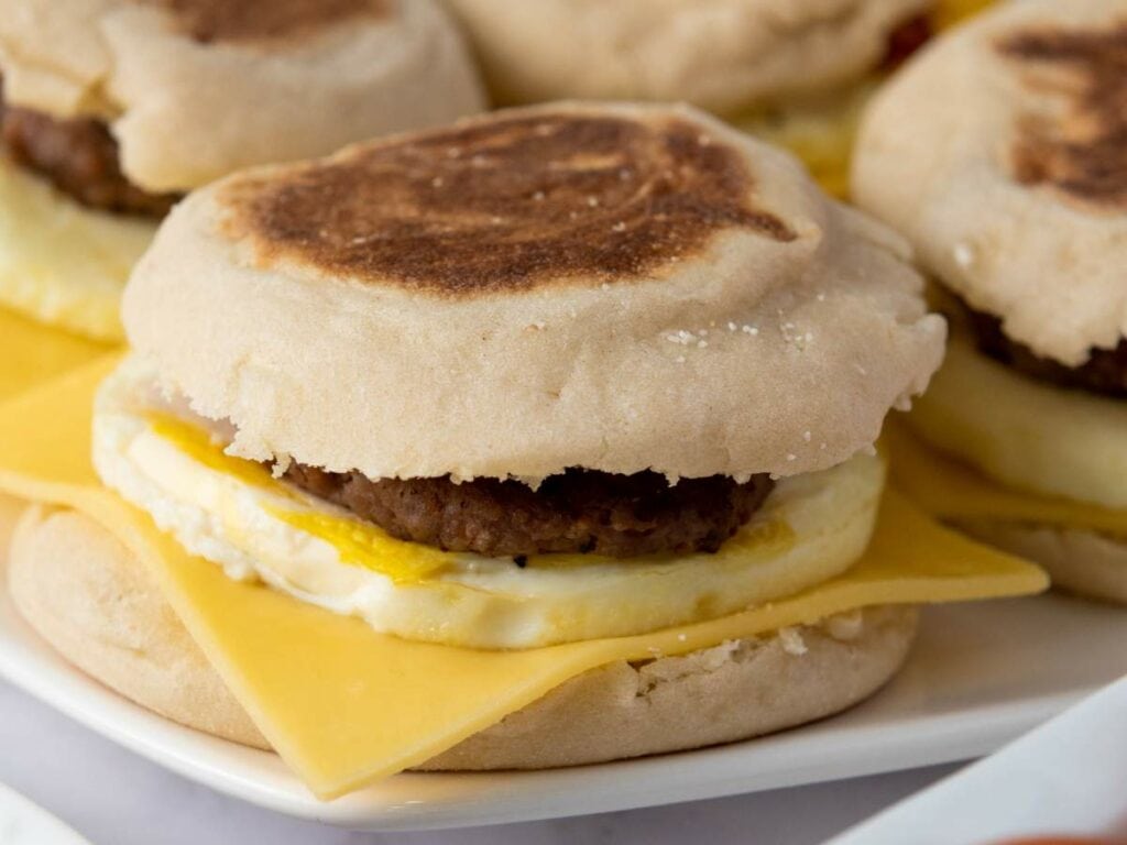 An English muffin breakfast sandwich with egg, a slice of cheddar cheese, and a sausage patty on a white plate.