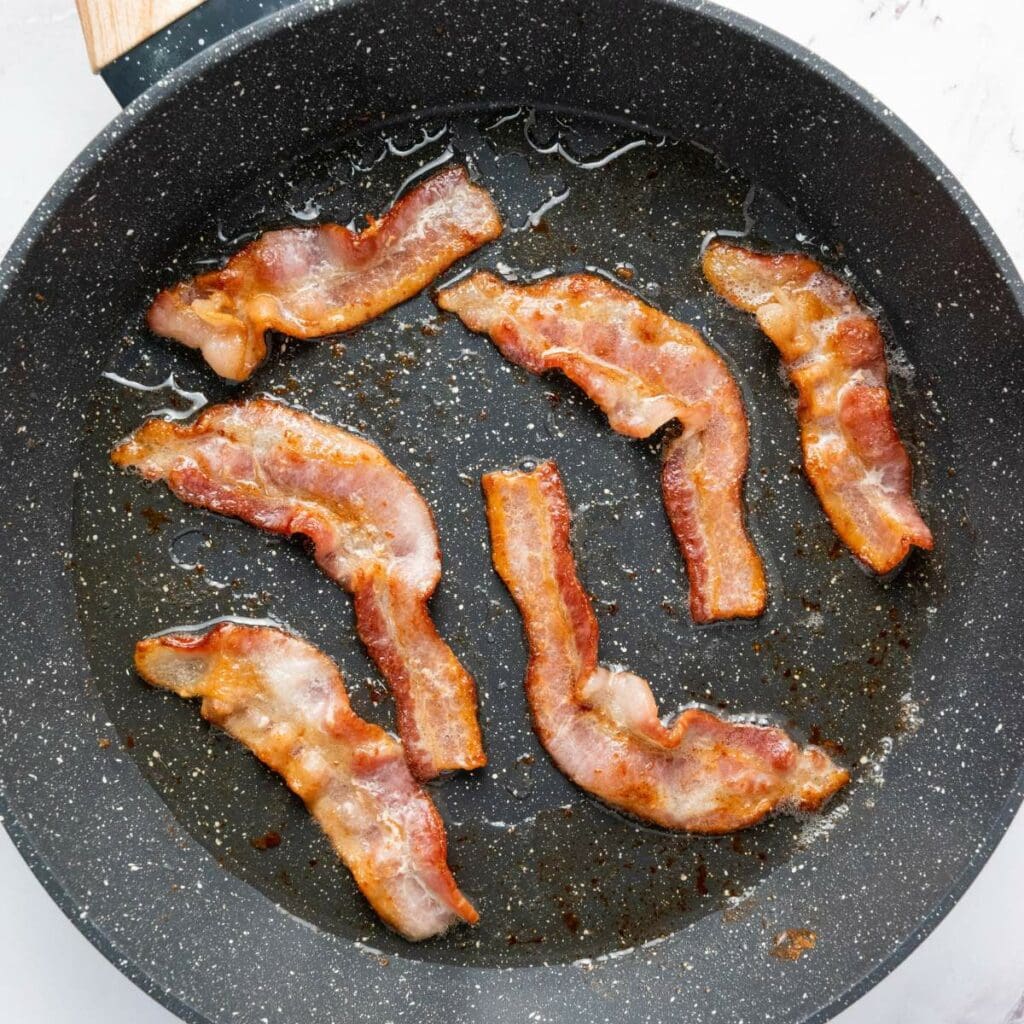 Six strips of cooked bacon in a frying pan with visible grease.