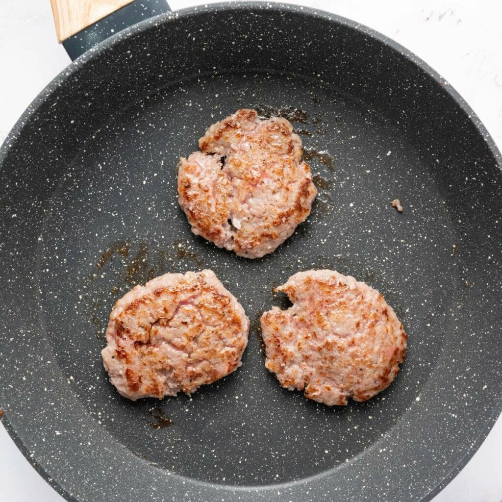 Three browned sausage patties are cooking in a nonstick frying pan.