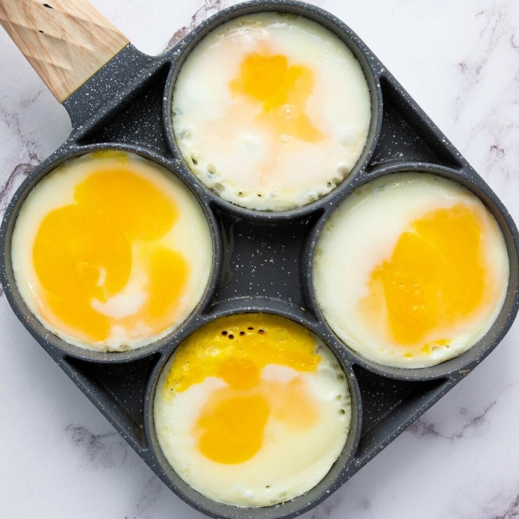 A square nonstick pan with four compartments, each holding a cooked sunny side up egg with runny yolks and set whites.