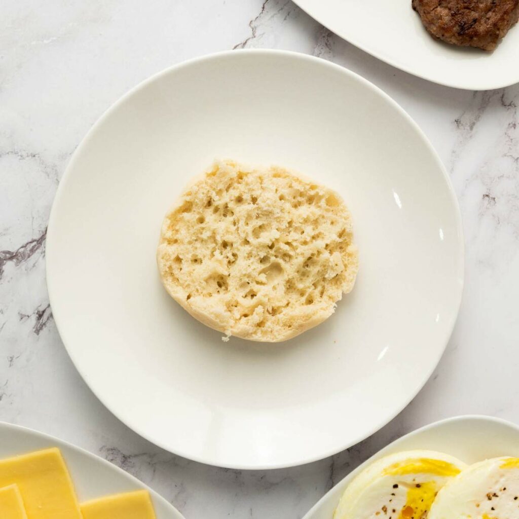 A plain, halved English muffin sits on a white plate, viewed from above on a marble surface. Slices of cheese, egg, and sausage are partially visible nearby.