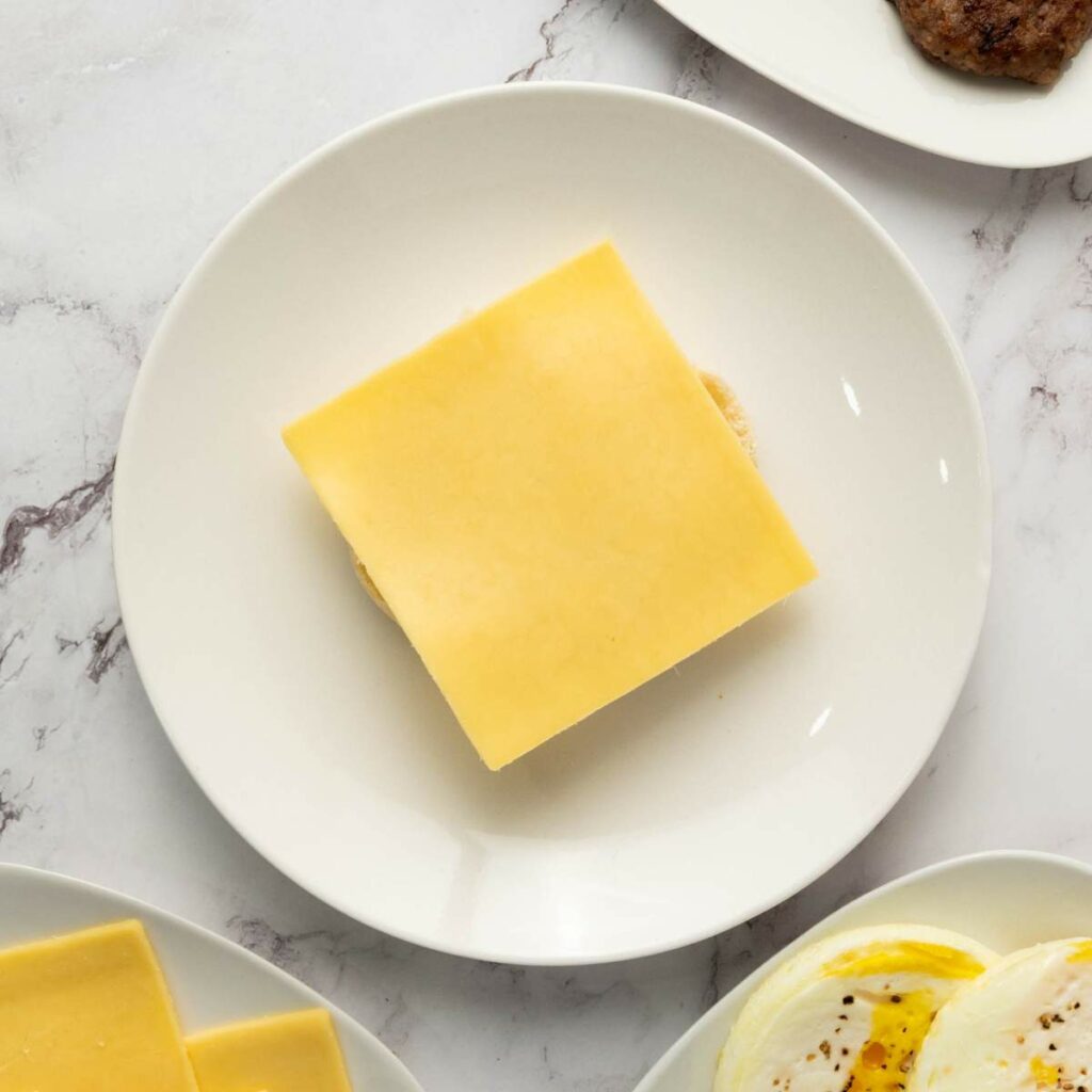 A slice of cheese on a plain hamburger bun sits centered on a white plate, with other food items partially visible around the edges.