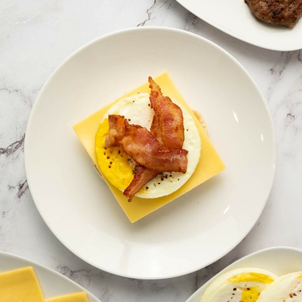 A slice of cheddar cheese, a cooked egg, and two strips of bacon are stacked on an English muffin half, served on a white plate.