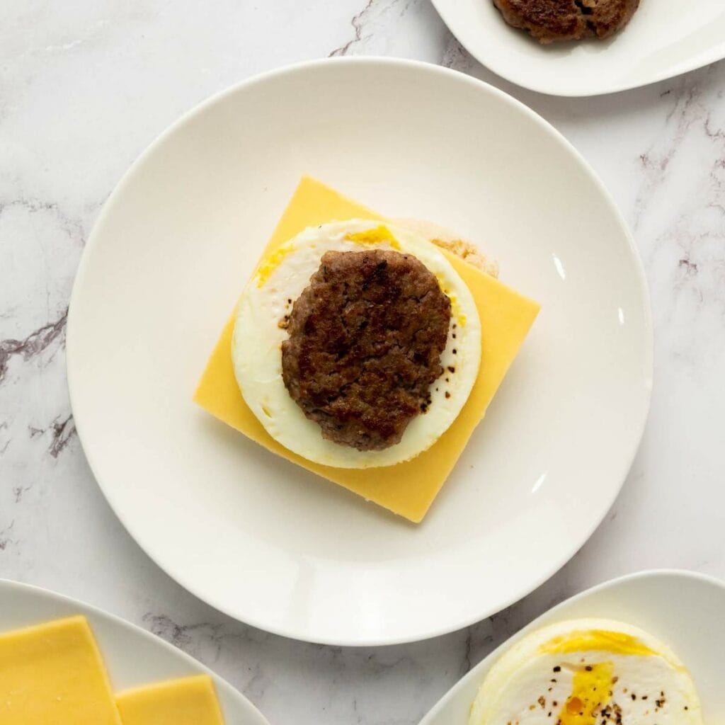 A breakfast sandwich with a slice of cheddar cheese, a cooked egg, and a sausage patty on a white plate, viewed from above.