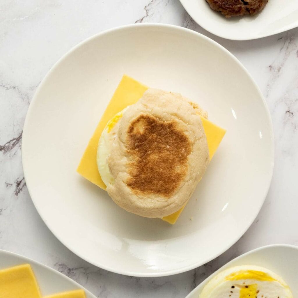 An English muffin breakfast sandwich with egg and cheddar cheese sits on a white plate, viewed from above.