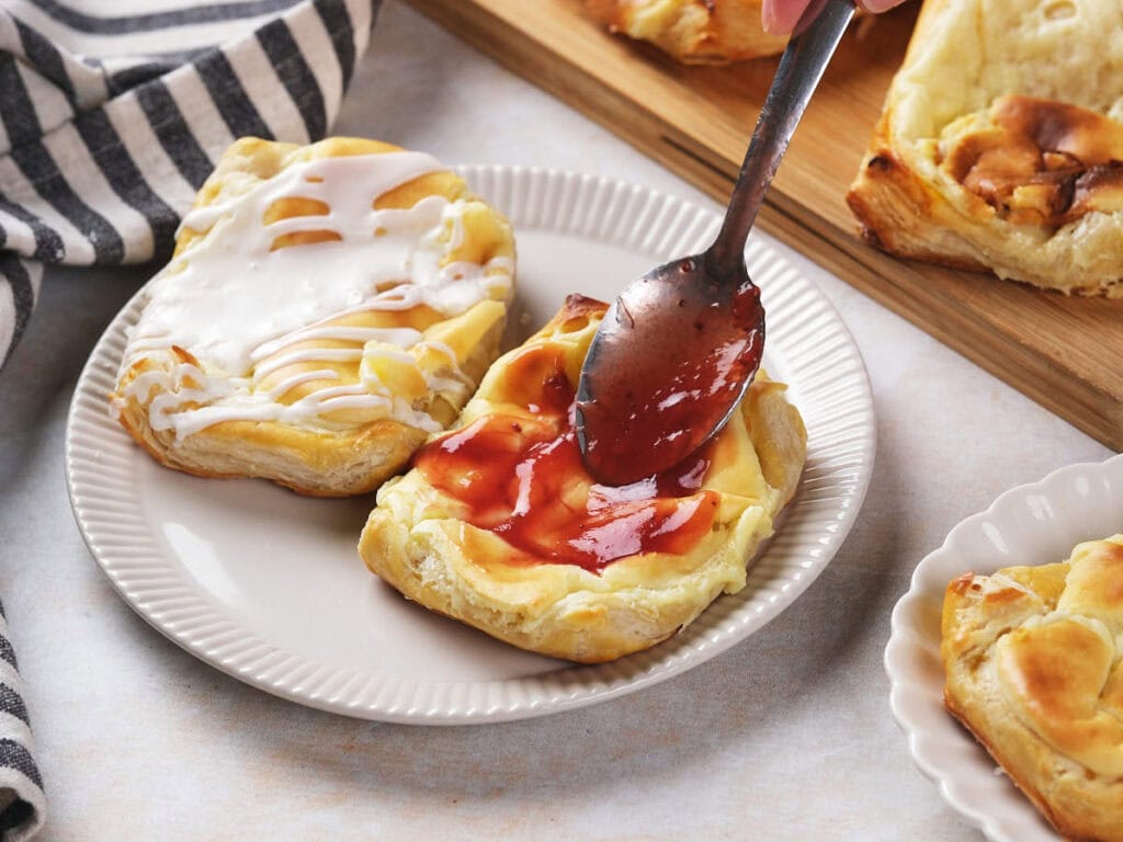 A hand spreads red jam on a cream cheese danish with a spoon, next to another pastry topped with white icing.