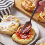 A hand spreads red fruit jam on a cream cheese danish with a spoon, next to another pastry topped with white icing.