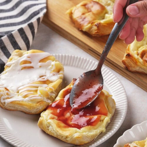 A hand spreads red fruit jam on a cream cheese danish with a spoon, next to another pastry topped with white icing.
