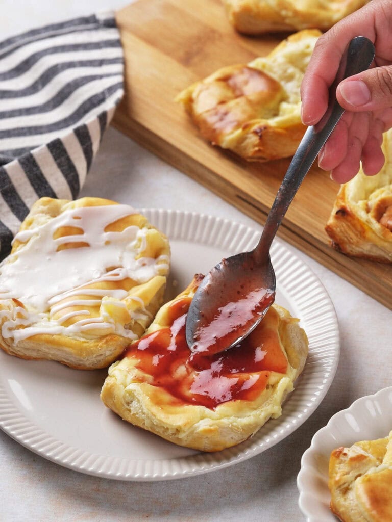 A hand spreads red fruit jam on a cream cheese danish with a spoon, next to another pastry topped with white icing.