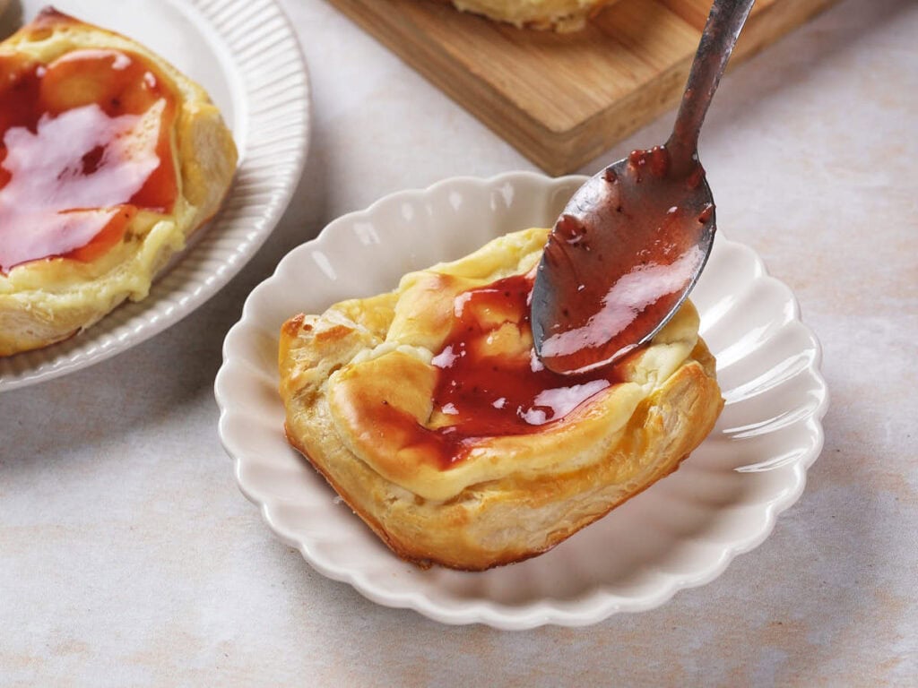 A spoon spreads red fruit jam over a cream cheese danish with a creamy filling, served on a white plate.
