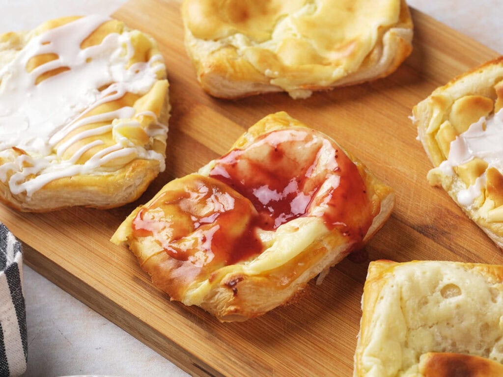 Cream cheese danish with flaky crusts and different toppings, including white icing and red fruit glaze.