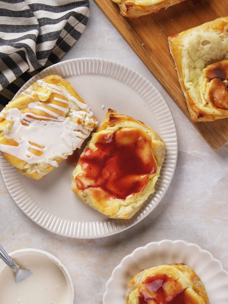 A plate with two pieces of danish pastry, one topped with white icing and the other with red fruit jam, next to a striped napkin and a wooden board with more pastries.