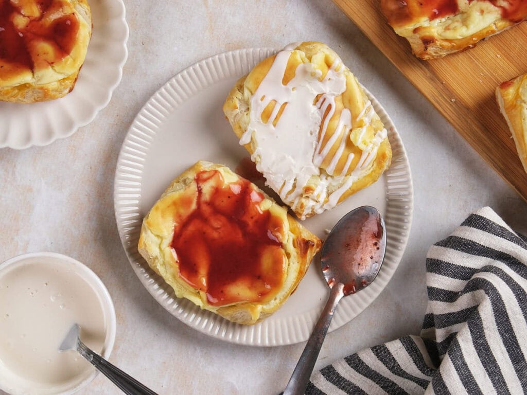 A plate with two cream cheese danishes, one topped with white icing and the other with red fruit glaze.