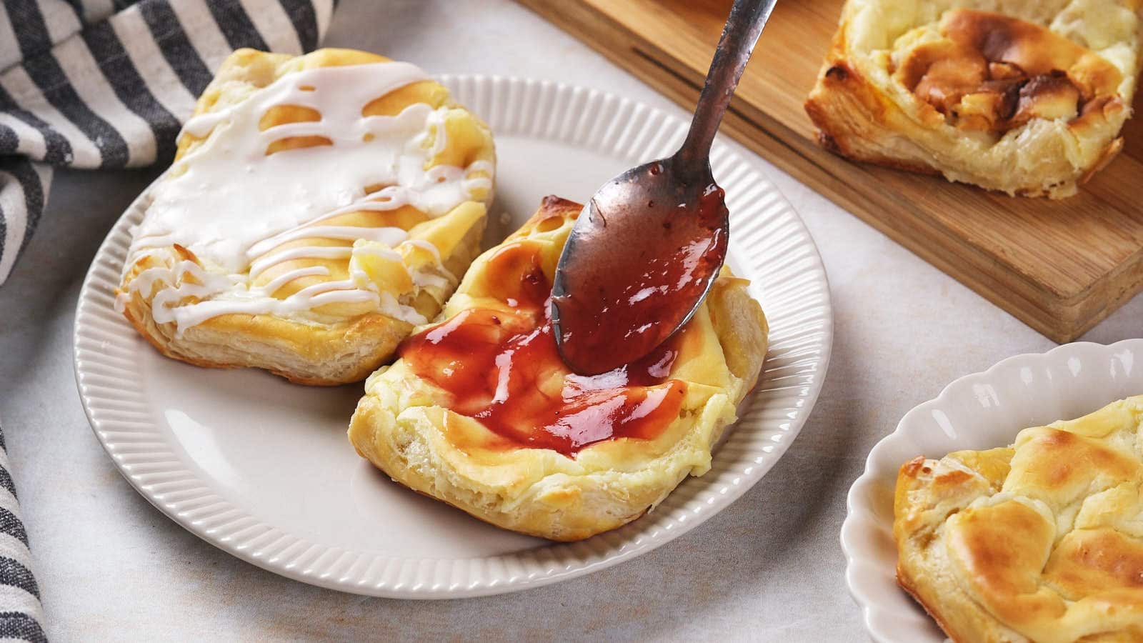 A spoon spreads red jam on a cream cheese danish with cream cheese filling.