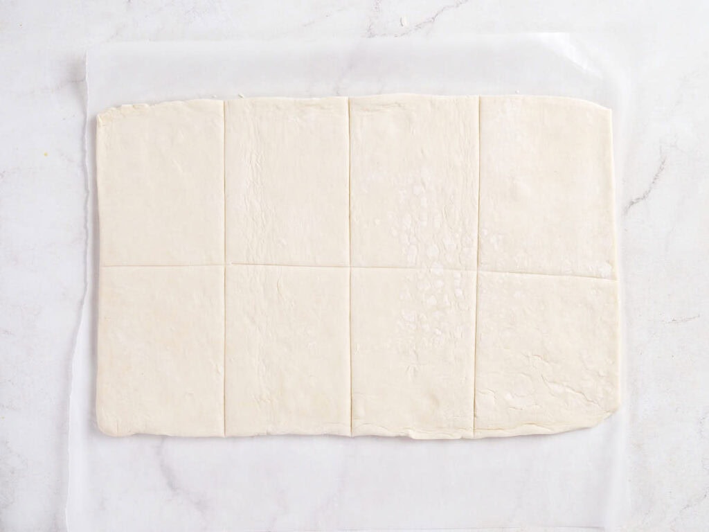 A sheet of puff pastry dough is laid flat on parchment paper and cut into eight equal rectangles.
