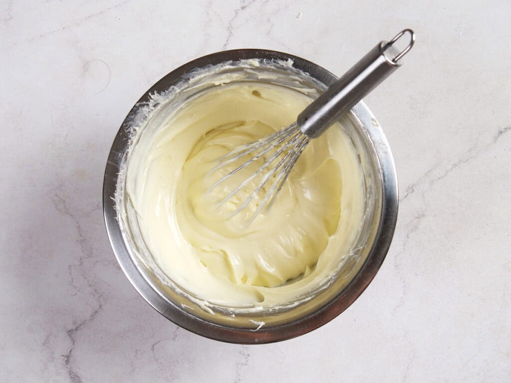 A metal whisk resting in a stainless steel bowl filled with smooth, creamy mixture on a light marble surface.