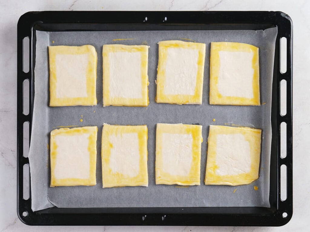 Eight rectangular pieces of puff pastry with egg wash on the borders are arranged on a parchment-lined baking tray.