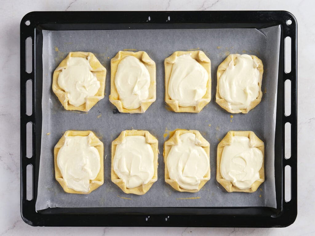Eight unbaked pastries with creamy white filling arranged on a baking tray lined with parchment paper, ready to be baked.