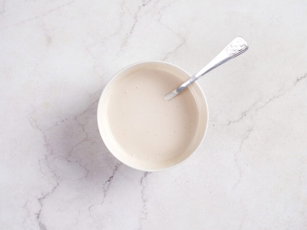 A bowl filled with a creamy white liquid and a metal spoon, placed on a light-colored marble surface.