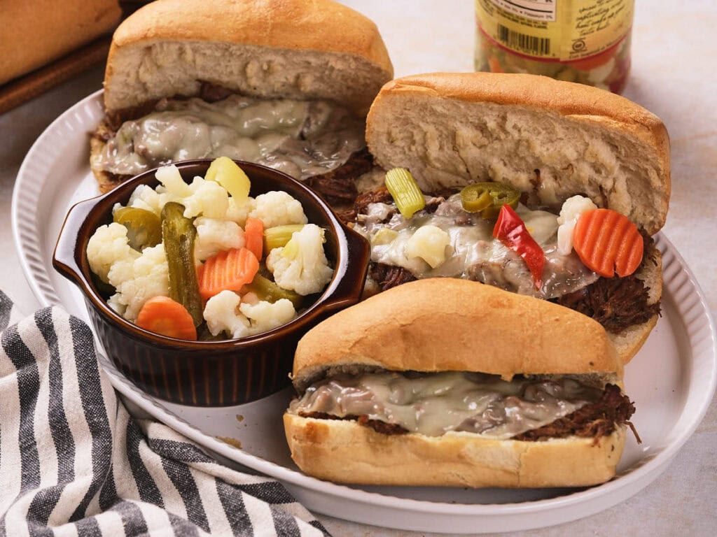 Three rItalian beef sandwiches with melted cheese on hoagie rolls are served on a white plate with a side of pickled vegetables in a small brown bowl.