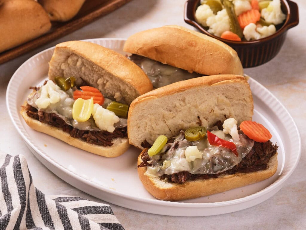 Three Italian beef sandwiches topped with melted cheese and pickled vegetables on a white plate, with a bowl of pickled vegetables in the background.