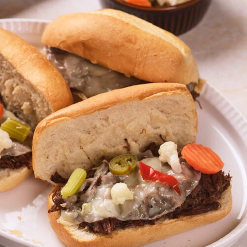 Three Italian beef sandwiches topped with melted cheese and pickled vegetables are served on a white plate.