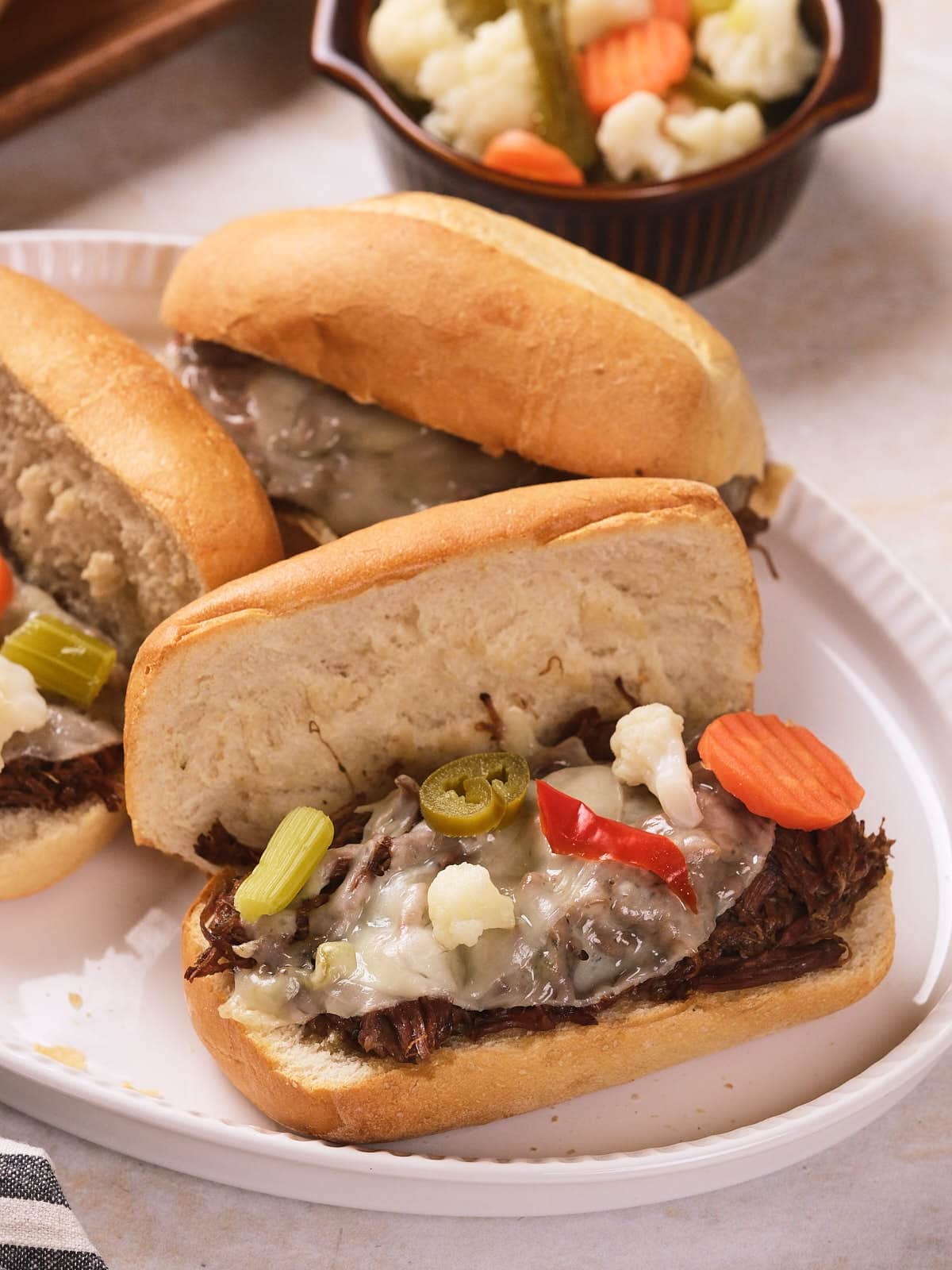 Three Italian beef sandwiches topped with melted cheese and pickled vegetables are served on a white plate.