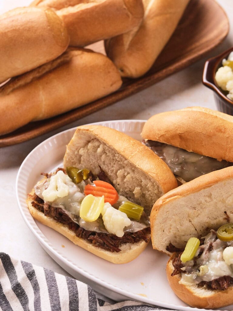 Three Italian beef sandwiches topped with melted cheese and pickled vegetables are served on a white plate.