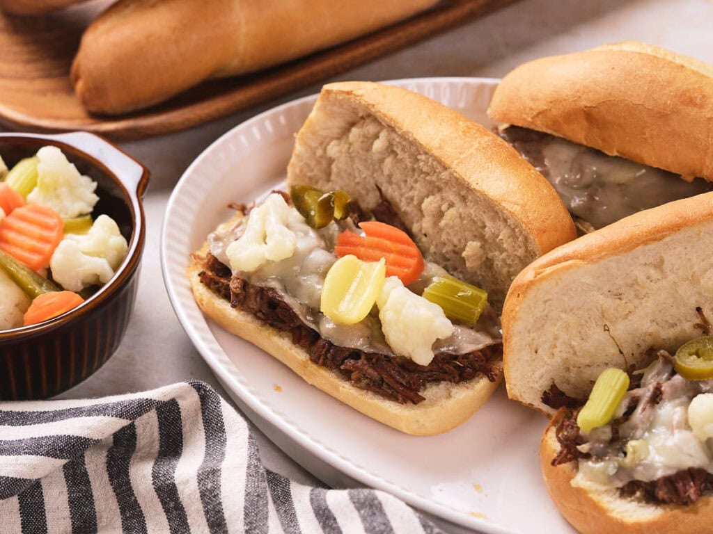 Three Italian beef sandwiches with melted cheese and pickled vegetables on a white plate, with extra pickled vegetables in a small bowl nearby.