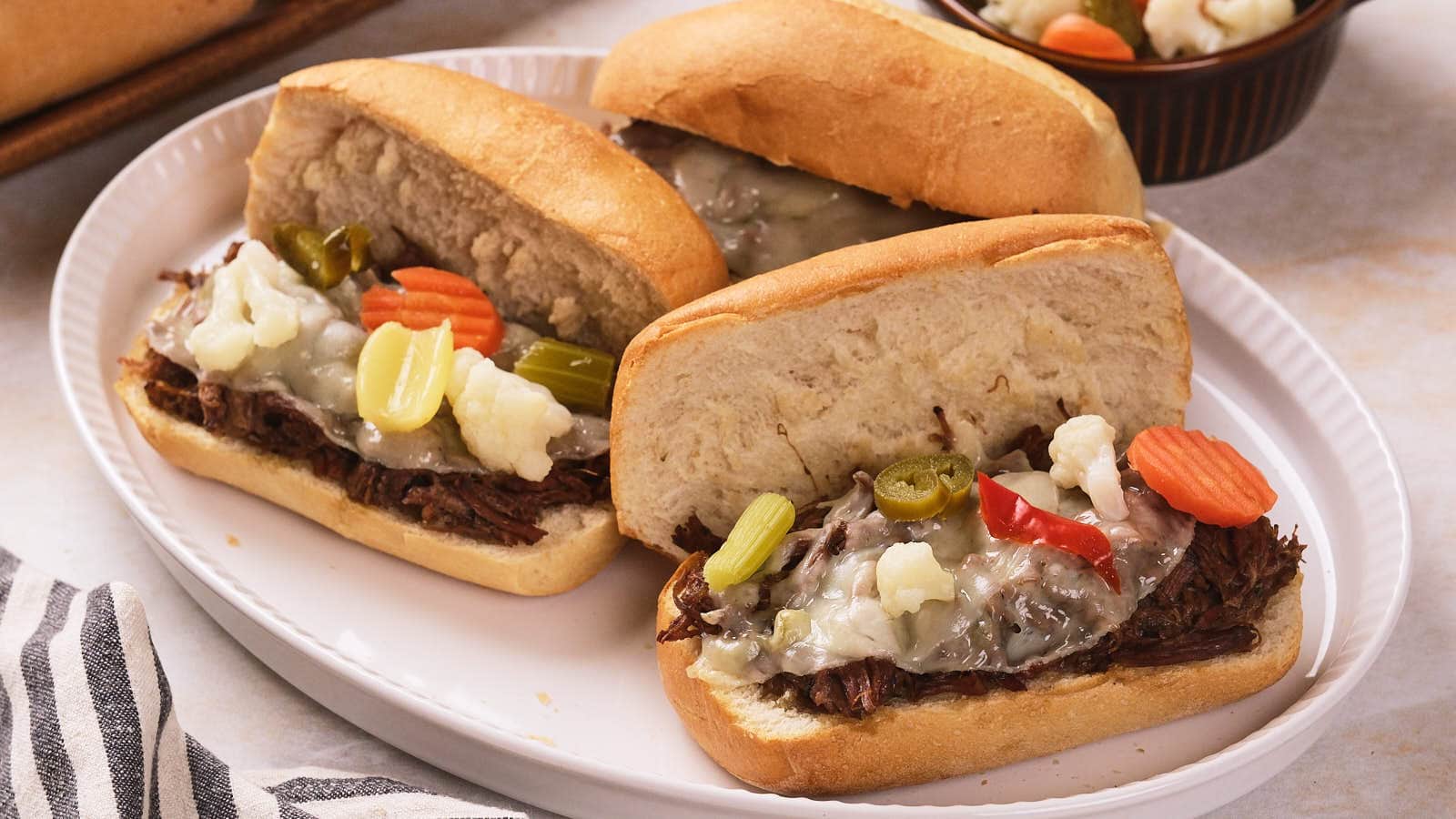 Three Italian beef sandwiches on hoagie rolls topped with melted cheese and pickled vegetables are served on a white plate.