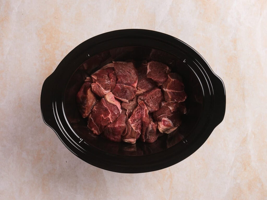 Chunks of raw beef placed inside a black slow cooker on a light-colored surface.