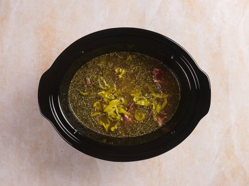 A black slow cooker filled with beef, sliced pepperoncini peppers, herbs, and broth, placed on a light-colored surface.