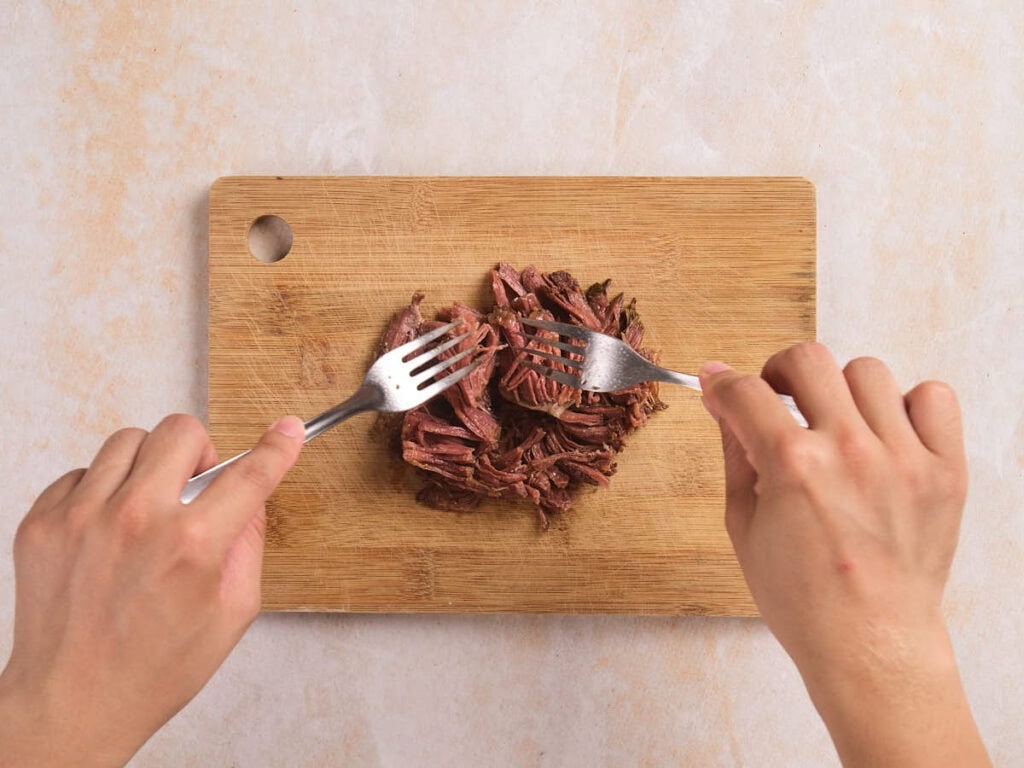 Two hands use forks to shred cooked meat on a wooden cutting board.