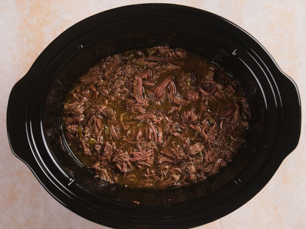 Shredded beef cooked in a dark broth inside a black slow cooker, viewed from above.