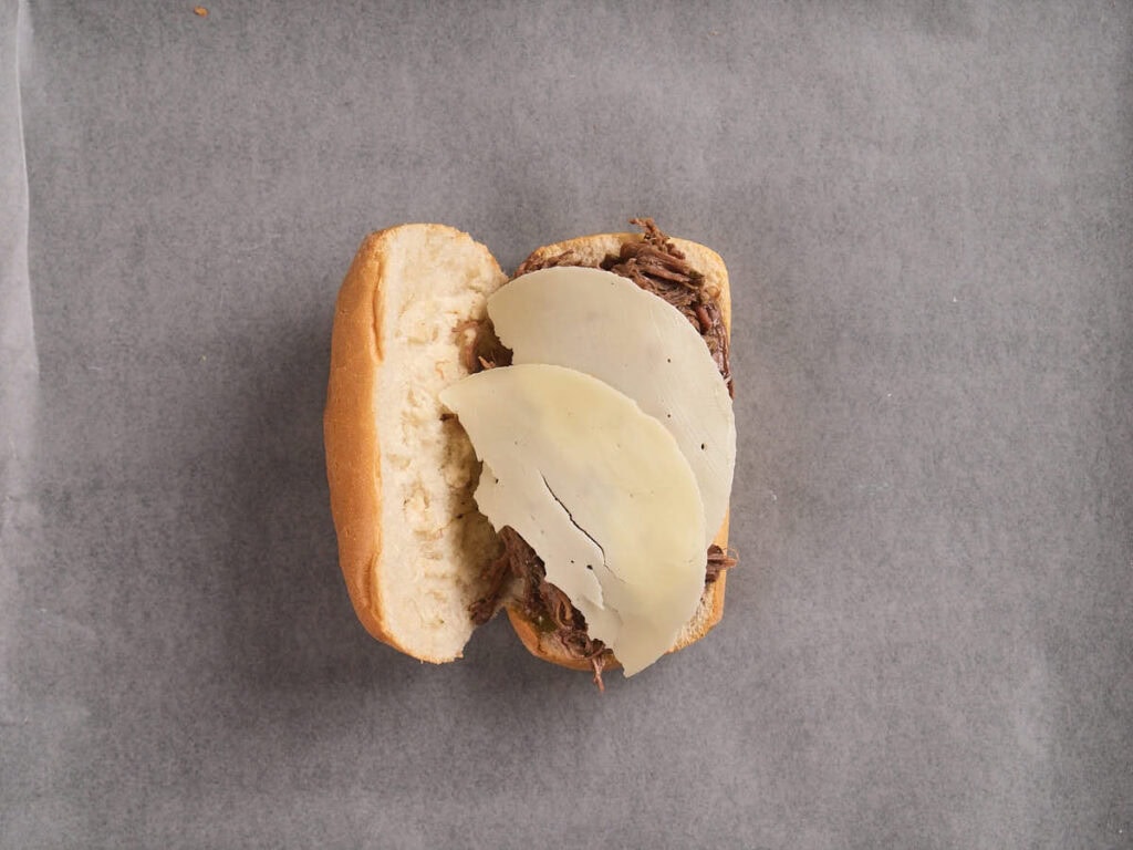 An open sandwich roll filled with shredded meat and topped with two slices of white cheese on a gray surface.