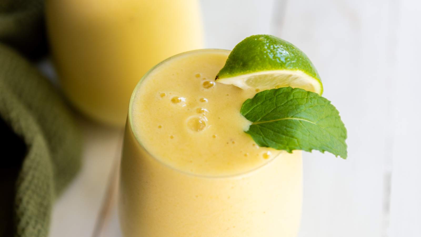 A glass pineapple smoothie garnished with a lime wedge and a mint leaf on a light-colored surface.