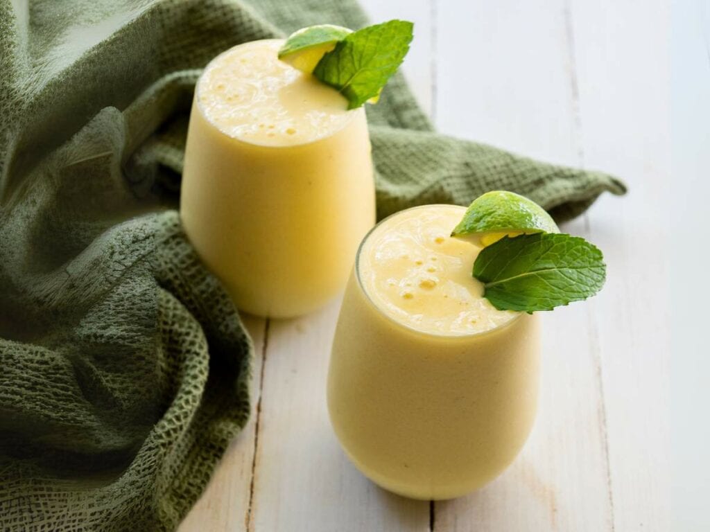 Two glasses of pineapple smoothie topped with fresh mint leaves are placed on a white wooden surface.