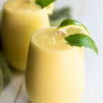 Two glasses of pineapple smoothie topped with lime wedges and mint leaves, placed on a light wooden surface.