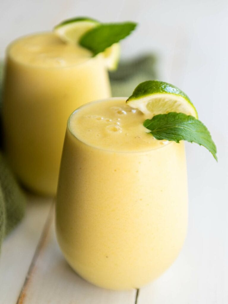 Two glasses of pineapple smoothie topped with lime wedges and mint leaves, placed on a light wooden surface.