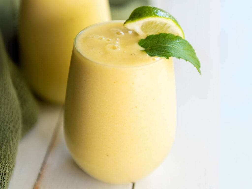 A glass of ypineapple smoothie garnished with a lime wedge and a mint leaf, placed on a white surface.