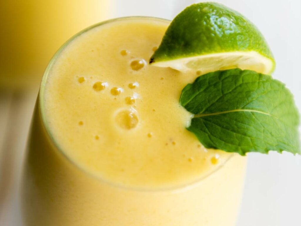 A close-up of a pineapple smoothie in a glass, garnished with a mint leaf and a slice of lime.
